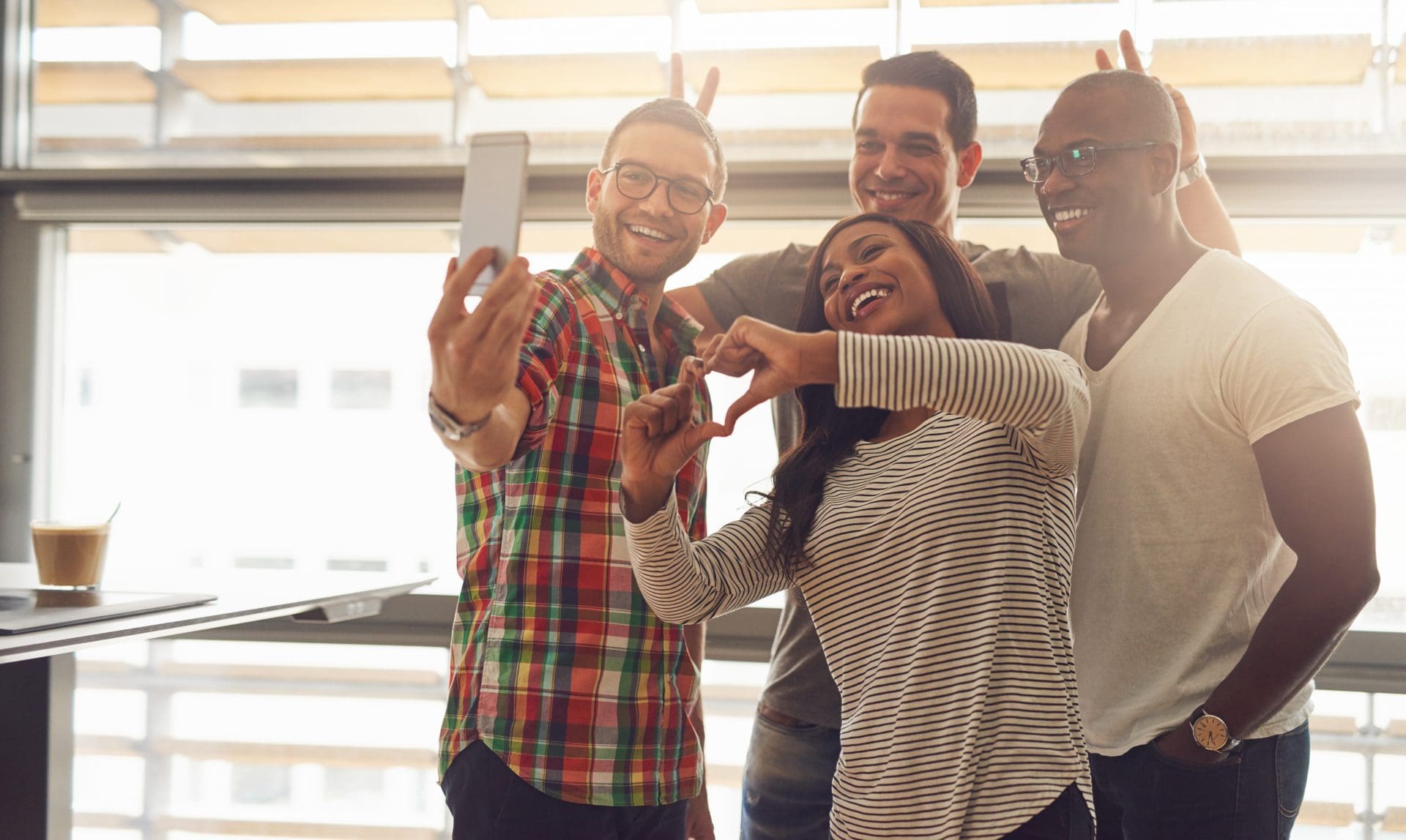 Group of four diverse silly employees taking self portrait and making symbolic hand gestures at small office in front of bright windows